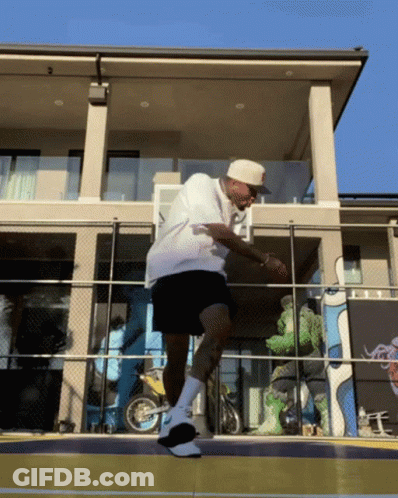 A Man In A White Shirt And Black Shorts Stands On A Basketball Court In Front Of A House GIF