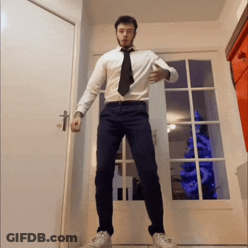A Man In A White Shirt And Black Tie Is Standing In Front Of A Christmas Tree GIF