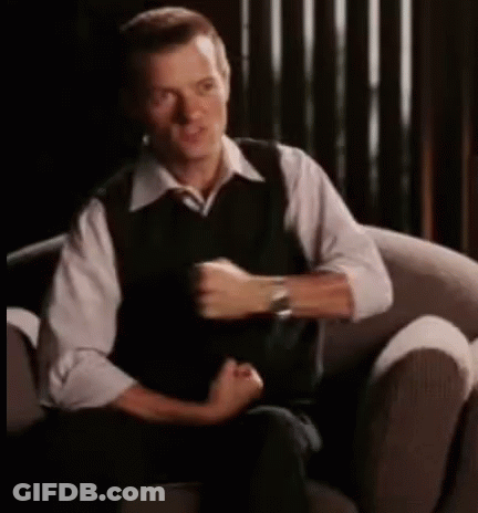 A Man In A White Shirt And Black Vest Is Sitting In A Chair . GIF