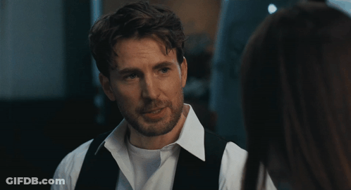 A Man In A White Shirt And Black Vest Looks At A Woman In A Dark Room GIF
