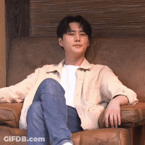 A Man In A White Shirt And Blue Jeans Is Sitting In A Chair GIF