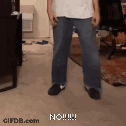 A Man In A White Shirt And Blue Jeans Is Standing In A Living Room With His Legs Crossed . GIF