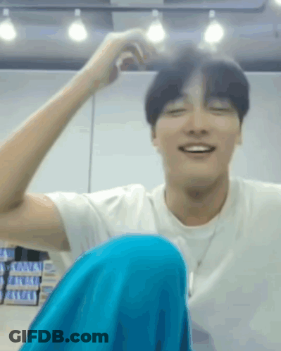 A Man In A White Shirt And Blue Pants Is Sitting On The Floor With His Legs Crossed And Smiling . GIF