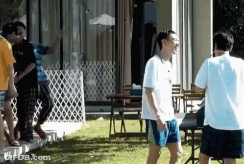 A Man In A White Shirt And Blue Shorts Is Standing In A Yard With Other People GIF