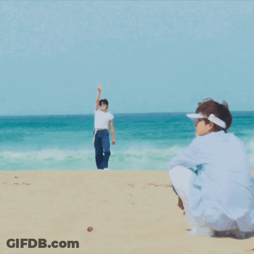 A Man In A White Shirt And Blue Shorts Stands On The Beach GIF