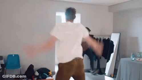A Man In A White Shirt And Brown Pants Is Dancing In A Room In Front Of A Mirror . GIF