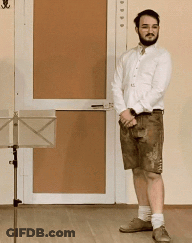 A Man In A White Shirt And Brown Shorts Stands In Front Of A Music Stand GIF