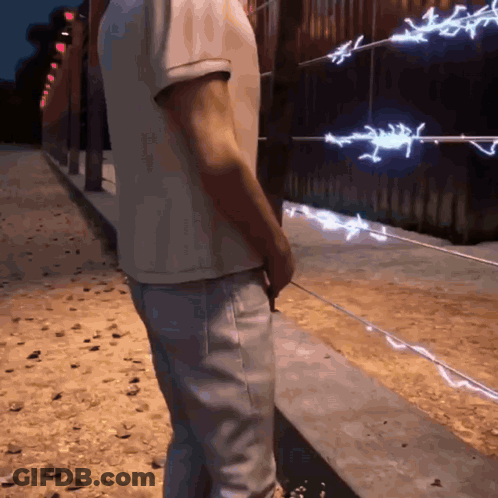 A Man In A White Shirt And Jeans Stands In Front Of A Barbed Wire Fence GIF