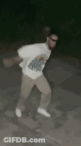 A Man In A White Shirt And Khaki Pants Is Dancing On A Sidewalk At Night . GIF