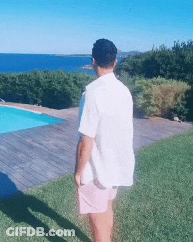 A Man In A White Shirt And Pink Shorts Is Standing In Front Of A Swimming Pool GIF