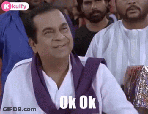 A Man In A White Shirt And Purple Scarf Is Sitting In A Crowd Of People And Smiling . GIF