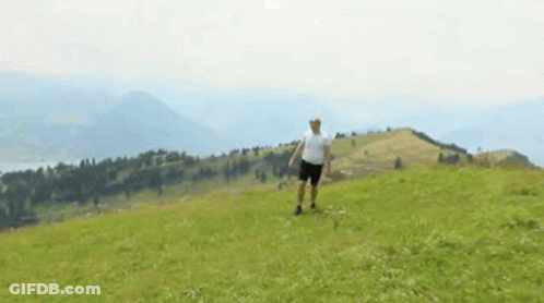 A Man In A White Shirt And Shorts Is Standing On Top Of A Grassy Hill . GIF