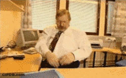 A Man In A White Shirt And Tie Sits At A Desk In Front Of A Computer Monitor GIF