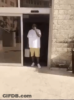 A Man In A White Shirt And Yellow Shorts Is Standing In A Doorway . GIF