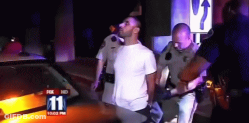 A Man In A White Shirt Is Being Escorted By Police Officers In Front Of A Fox 11 News Car GIF