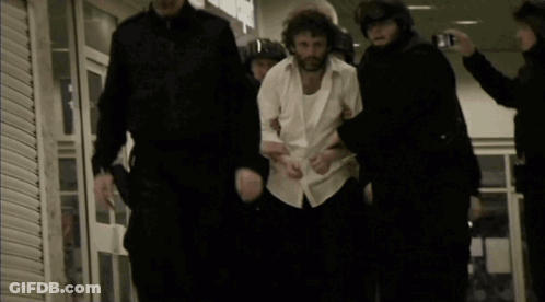 A Man In A White Shirt Is Being Led By A Group Of Police Officers GIF