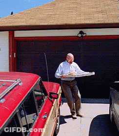 A Man In A White Shirt Is Carrying A Pizza In Front Of A Garage Door GIF