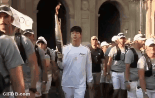 A Man In A White Shirt Is Carrying A Torch In Front Of A Crowd Of People . GIF