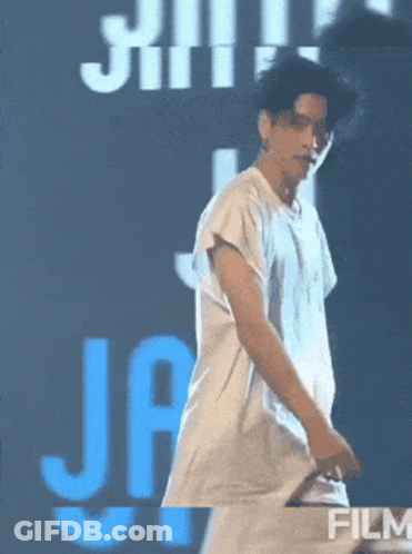 A Man In A White Shirt Is Dancing In Front Of A Blue Sign That Says Film GIF
