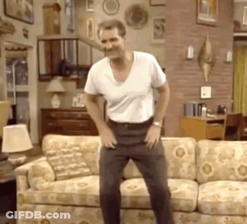 A Man In A White Shirt Is Dancing In Front Of A Couch In A Living Room GIF