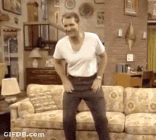 A Man In A White Shirt Is Dancing In Front Of A Couch . GIF