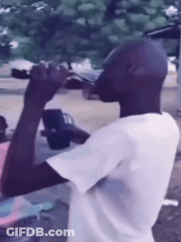 A Man In A White Shirt Is Drinking From A Bottle Outside GIF