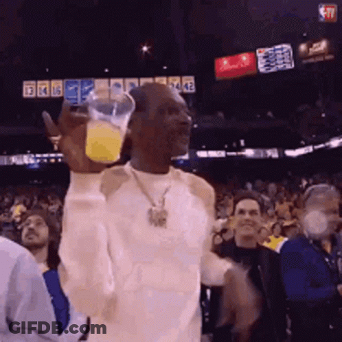 A Man In A White Shirt Is Holding A Glass Of Orange Juice In His Hand GIF