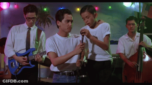 A Man In A White Shirt Is Holding A Microphone While Another Man Plays A Guitar GIF