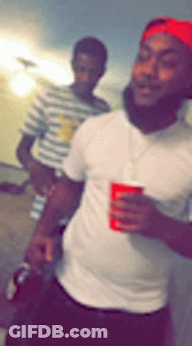 A Man In A White Shirt Is Holding A Red Cup While Another Man Stands Behind Him . GIF