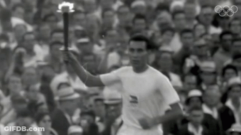 A Man In A White Shirt Is Holding A Torch In Front Of A Crowd With The Olympic Rings In The Background GIF
