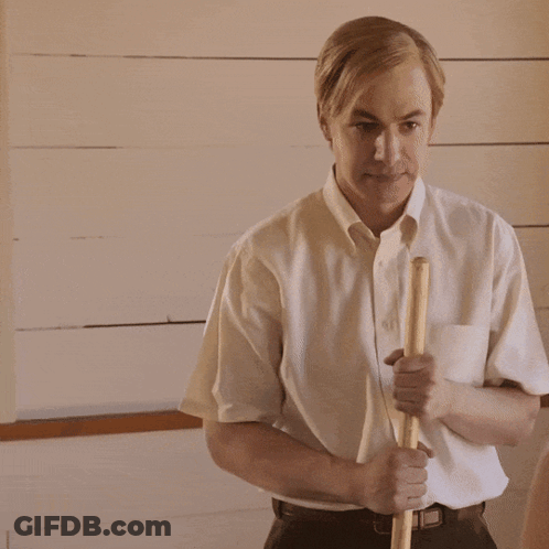 A Man In A White Shirt Is Holding A Wooden Stick In His Right Hand GIF