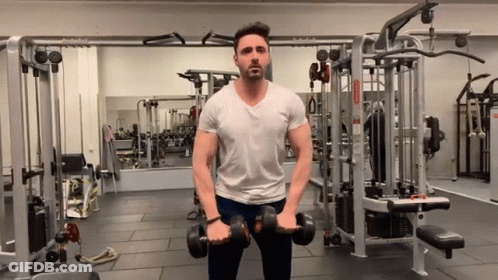 A Man In A White Shirt Is Holding Dumbbells In A Gym . GIF