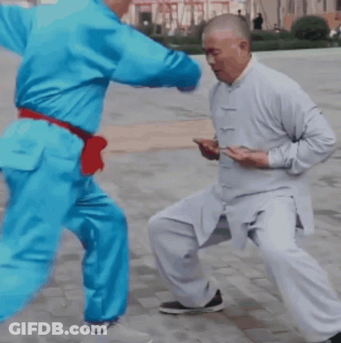 A Man In A White Shirt Is Kicking Another Man In A Blue Outfit GIF