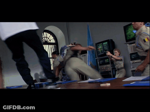 A Man In A White Shirt Is Kicking Another Man In A Room With Other People GIF
