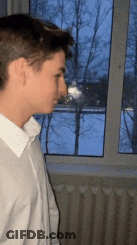 A Man In A White Shirt Is Looking Out A Window GIF