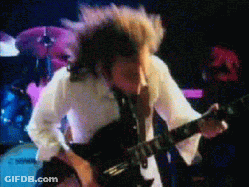 A Man In A White Shirt Is Playing A Guitar In Front Of A Drum Set GIF
