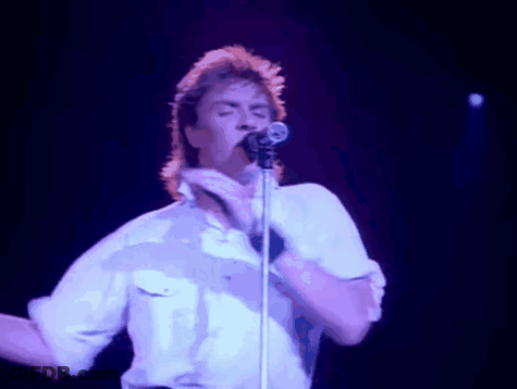 A Man In A White Shirt Is Singing Into A Microphone And Making A Peace Sign GIF