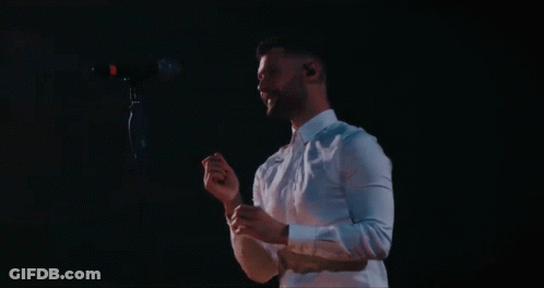 A Man In A White Shirt Is Singing Into A Microphone In A Dark Room GIF