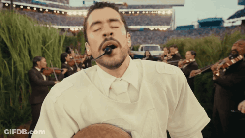 A Man In A White Shirt Is Singing Into A Microphone In Front Of A Band GIF
