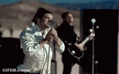 A Man In A White Shirt Is Singing Into A Microphone While Another Man Plays A Guitar . GIF