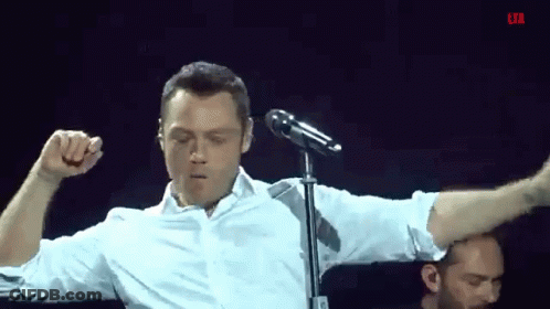 A Man In A White Shirt Is Singing Into A Microphone While Flexing His Muscles GIF