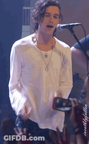 A Man In A White Shirt Is Singing Into A Microphone While Holding A Camera In Front Of A Crowd GIF