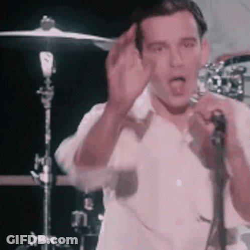 A Man In A White Shirt Is Singing Into A Microphone While Standing In Front Of A Drum Set . GIF