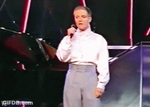 A Man In A White Shirt Is Singing Into A Microphone While Standing On A Stage GIF