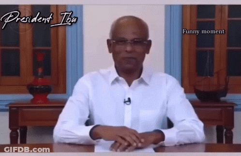 A Man In A White Shirt Is Sitting At A Desk With A Funny Moment Written On The Bottom GIF