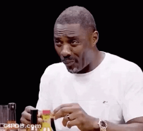 A Man In A White Shirt Is Sitting At A Table With A Bottle Of Hot Sauce In His Hand . GIF