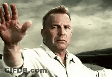 A Man In A White Shirt Is Sitting In A Car Waving His Hand . GIF