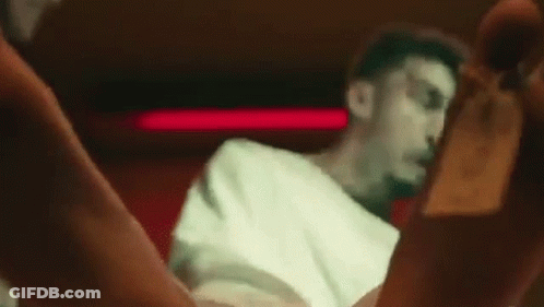 A Man In A White Shirt Is Sitting In A Chair In A Dark Room With His Legs Crossed . GIF