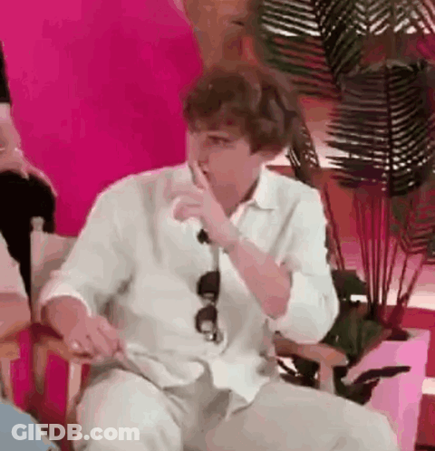 A Man In A White Shirt Is Sitting In A Chair With His Hand To His Mouth . GIF