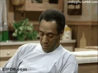 A Man In A White Shirt Is Sitting In A Kitchen Looking Down . GIF